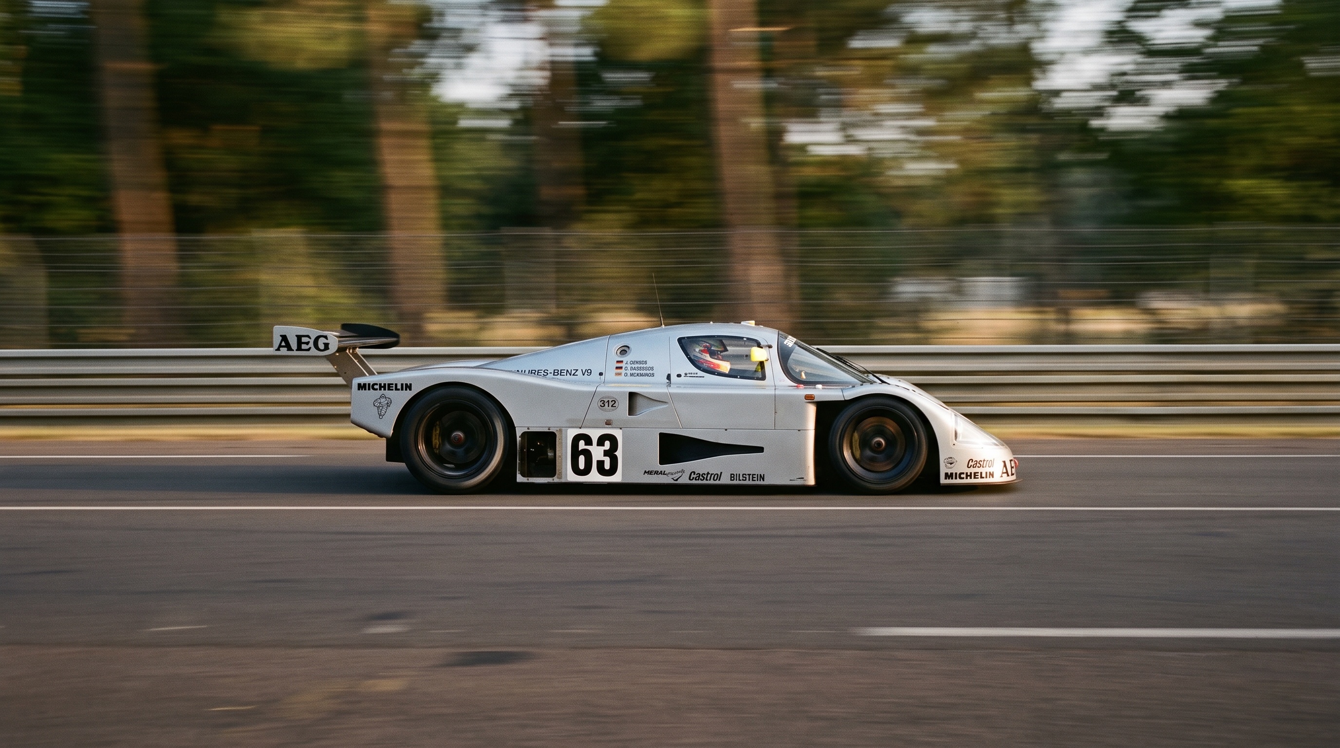 Srebrny Sauber-Mercedes C9 z numerem 63 pędzący z pełną prędkością obok barierek i drzew